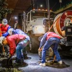 Trabalhadores fazem a limpeza de um sistema de drenagem urbana à noite, sob um viaduto, utilizando equipamentos e maquinaria.