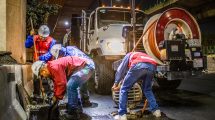 Trabalhadores fazem a limpeza de um sistema de drenagem urbana à noite, sob um viaduto, utilizando equipamentos e maquinaria.