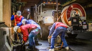 Trabalhadores fazem a limpeza de um sistema de drenagem urbana à noite, sob um viaduto, utilizando equipamentos e maquinaria.
