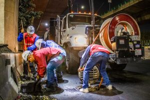 Trabalhadores fazem a limpeza de um sistema de drenagem urbana à noite, sob um viaduto, utilizando equipamentos e maquinaria.