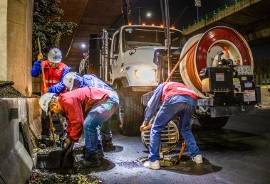 Trabalhadores fazem a limpeza de um sistema de drenagem urbana à noite, sob um viaduto, utilizando equipamentos e maquinaria.