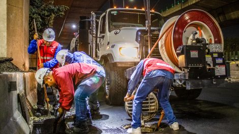 Trabalhadores fazem a limpeza de um sistema de drenagem urbana à noite, sob um viaduto, utilizando equipamentos e maquinaria.