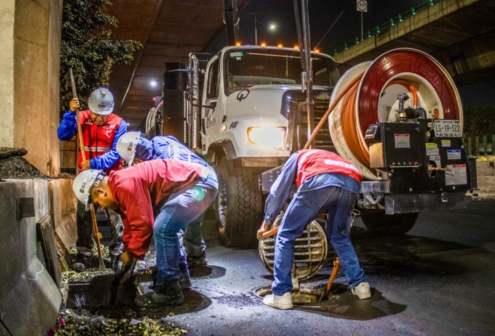 Trabalhadores fazem a limpeza de um sistema de drenagem urbana à noite, sob um viaduto, utilizando equipamentos e maquinaria.