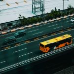 Ônibus urbano amarelo circula por uma avenida movimentada entre carros, visto de cima.