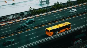 Ônibus urbano amarelo circula por uma avenida movimentada entre carros, visto de cima.