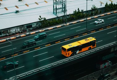 Ônibus urbano amarelo circula por uma avenida movimentada entre carros, visto de cima.