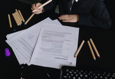 Profissional analisa documentos sobre uma mesa de trabalho, com lápis coloridos ao lado.