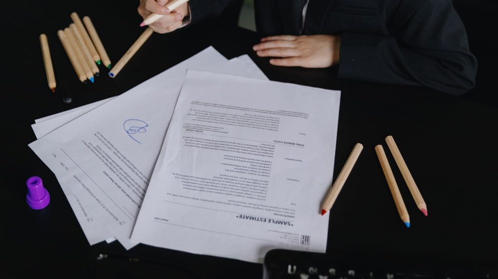 Profissional analisa documentos sobre uma mesa de trabalho, com lápis coloridos ao lado.