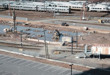 Vista aérea de um canteiro de obras ferroviárias ao lado de trilhos, com trabalhadores e máquinas em operação.