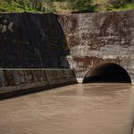 Estrutura de drenagem pluvial de concreto, com água escura escoando para um túnel entre paredes cobertas de musgo.