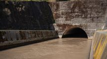 Estrutura de drenagem pluvial de concreto, com água escura escoando para um túnel entre paredes cobertas de musgo.