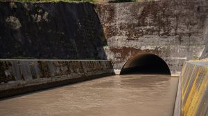 Estrutura de drenagem pluvial de concreto, com água escura escoando para um túnel entre paredes cobertas de musgo.