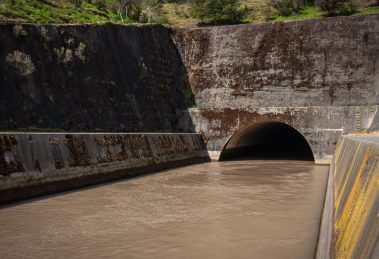 Estrutura de drenagem pluvial de concreto, com água escura escoando para um túnel entre paredes cobertas de musgo.