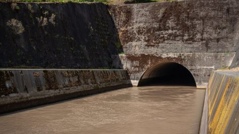 Estrutura de drenagem pluvial de concreto, com água escura escoando para um túnel entre paredes cobertas de musgo.