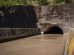 Estrutura de drenagem pluvial de concreto, com água escura escoando para um túnel entre paredes cobertas de musgo.