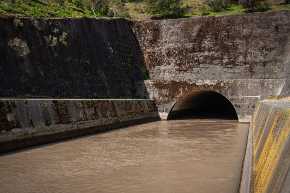 Estrutura de drenagem pluvial de concreto, com água escura escoando para um túnel entre paredes cobertas de musgo.
