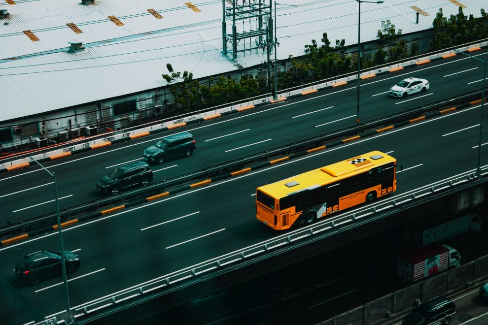 Ônibus urbano amarelo circula por uma avenida movimentada entre carros, visto de cima.
