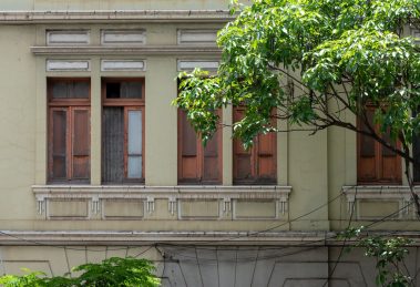 Fachada de uma construção histórica com janelas de madeira, cercada por vegetação em Belo Horizonte.